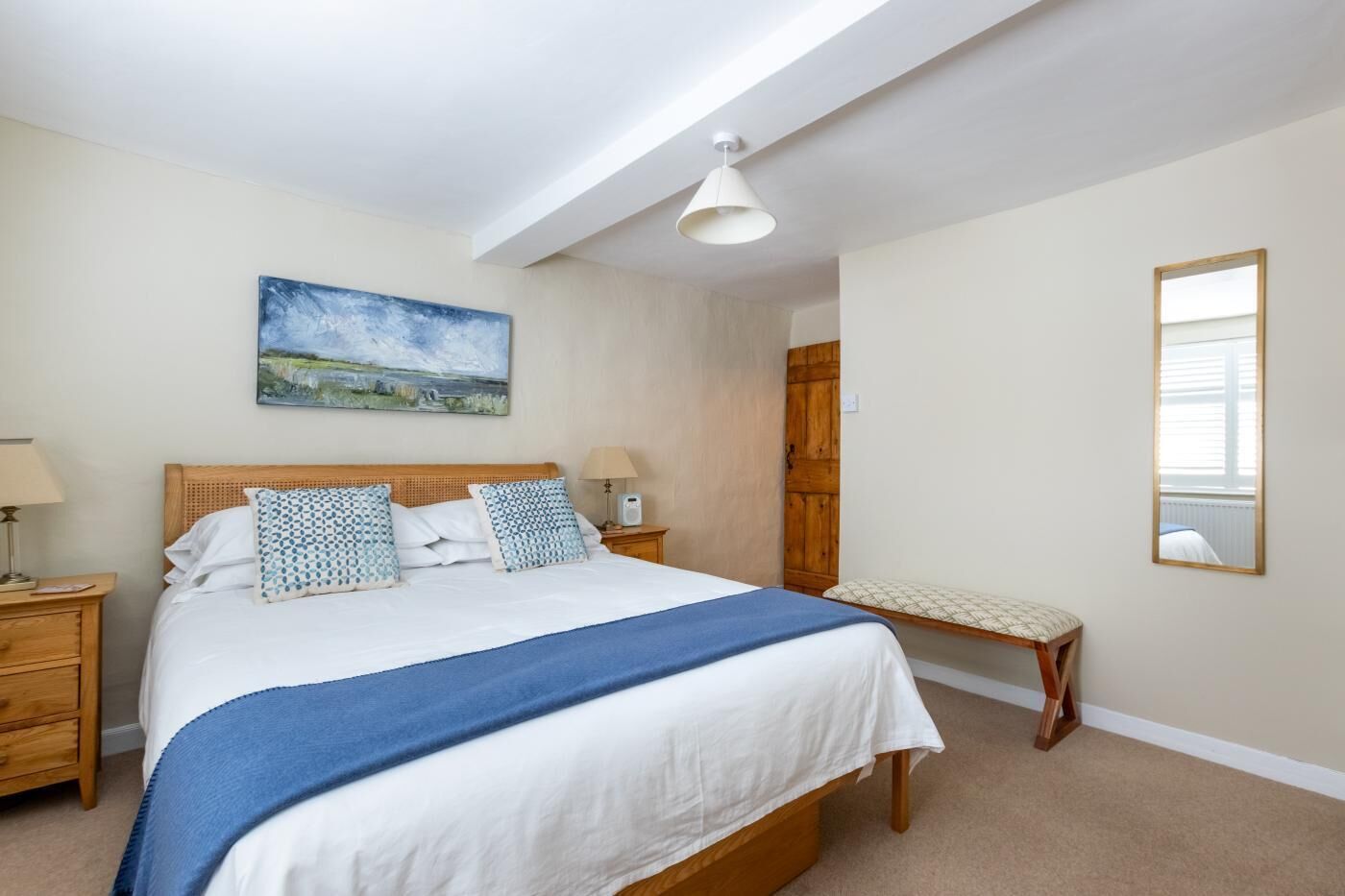 A bedroom with a large bed, a wooden headboard, a bench, a bedside table with a lamp, a wall-mounted painting, and a mirror.