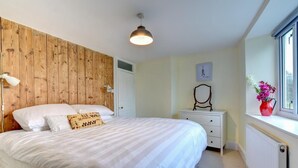 Una camera da letto con una testiera in legno, un letto con biancheria da letto bianca, una lampada da comodino, un comò, uno specchio e una finestra con un vaso di fiori.