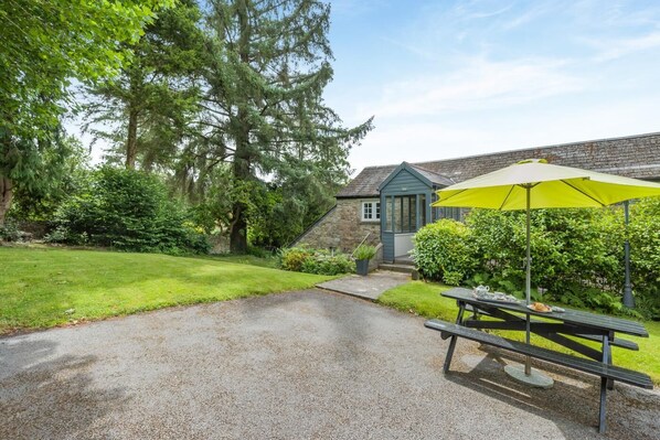 A gravel pathway leading to a stone building with a blue door, a picnic table with a yellow umbrella, and lush greenery. - 1 Bed in Two Waters Foot (oc-a27332) (Warleggan)