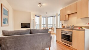 Un appartement moderne avec un canapé gris, une cuisine dotée de meubles en bois, un coin repas avec une table et des chaises, et un téléviseur fixé au mur.