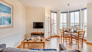 Un salon comprenant un canapé, une table basse, un téléviseur et une bibliothèque. Il y a également un balcon avec une table à manger et des chaises.