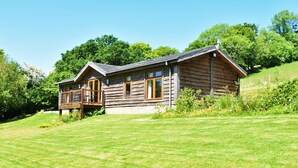A wooden cabin with a balcony, surrounded by a grassy lawn and trees.