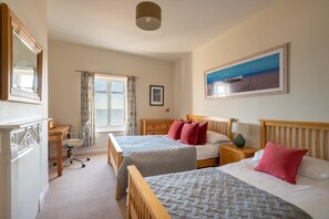 Une chambre d’hôtel avec deux lits, un bureau, une chaise, une fenêtre avec des rideaux, un tableau au mur et une lampe.