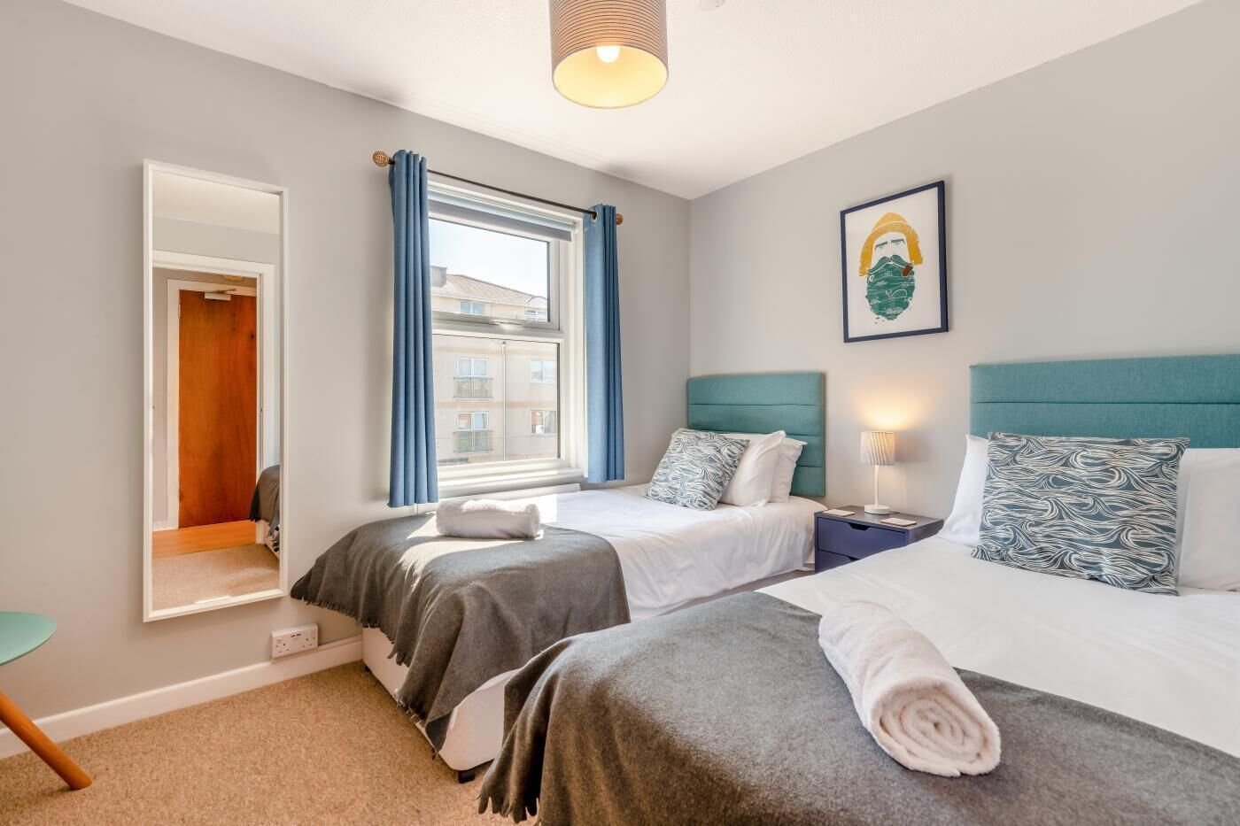 A hotel room with two beds, a window with curtains, a bedside table with a lamp, and a framed picture on the wall.