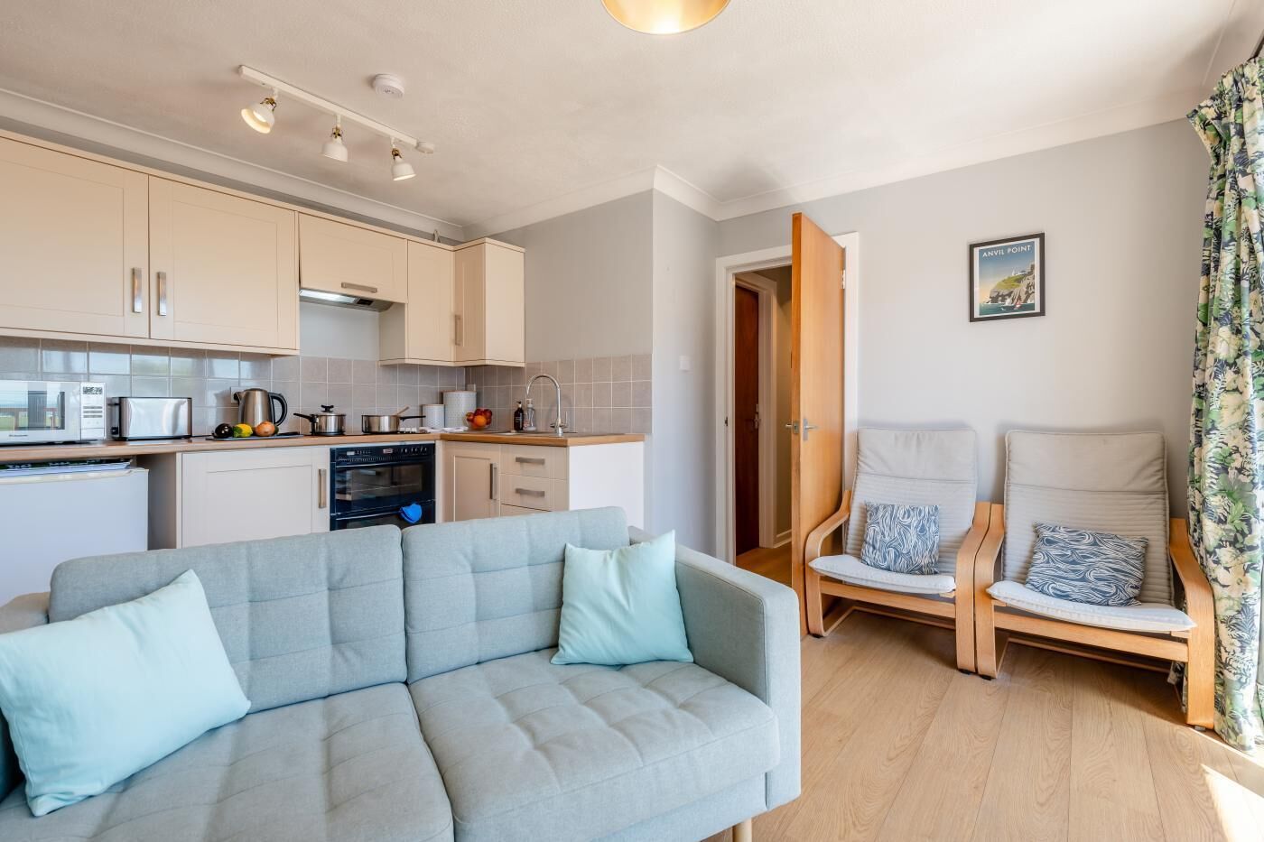 A modern kitchen with a built-in oven, microwave, and toaster. There is a grey sofa with light blue cushions in the living area, and two wooden chairs with patterned cushions.