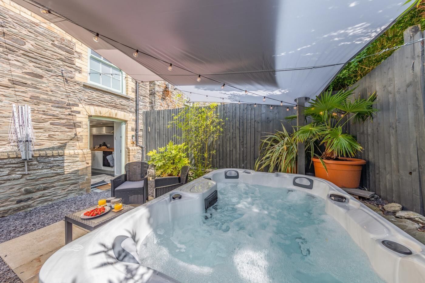 A hot tub in a backyard with stone walls, a wooden fence, and potted plants.