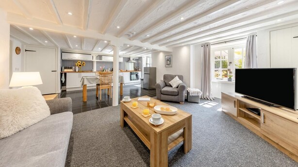 Ein modernes Wohnzimmer mit Sofa, Couchtisch und Fernseher. Im Hintergrund ist eine Küche mit Essbereich zu sehen.