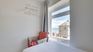 Ein Schlafzimmer mit einem Bett, einem Sessel mit floralem Bezug, einem Kleiderständer mit bunten Pinnwänden und Blick auf ein Gebäude durch ein Fenster.