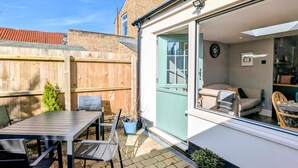 Un patio con una mesa y sillas, una cerca de madera y una puerta azul que da a una habitación con un sofá y un reloj.