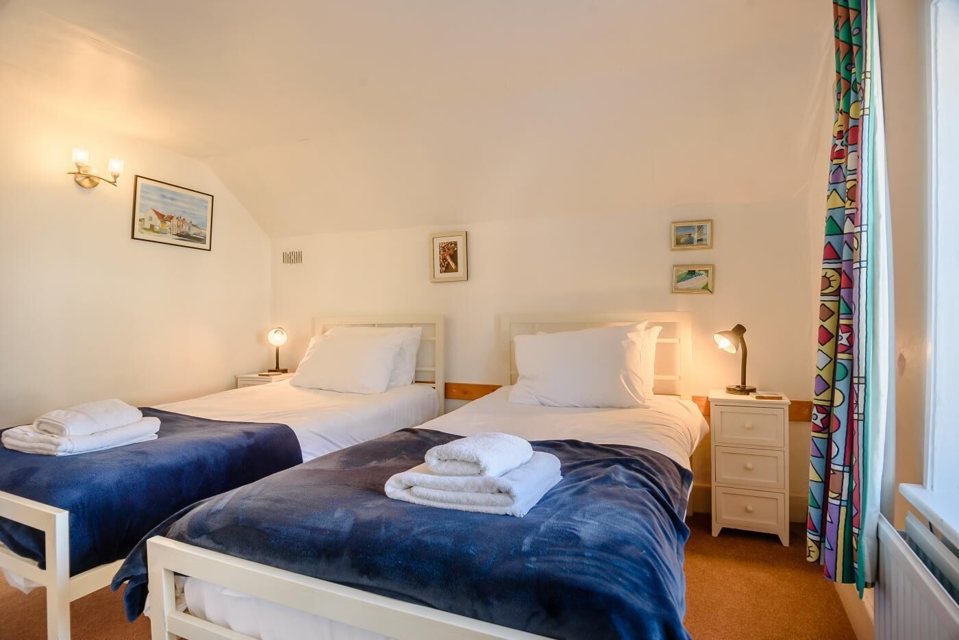 A hotel room with two beds, each with a white headboard, a bedside table with a lamp, and a small drawer. There are two wall-mounted light fixtures and a framed picture above each bed.
