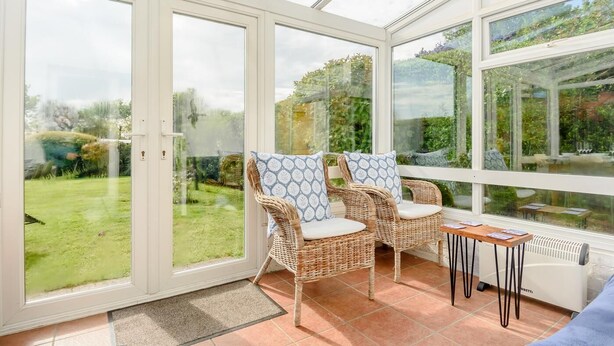 Een oranjerie met rieten stoelen en een tafeltje, grote glazen deuren en uitzicht op een tuin.
