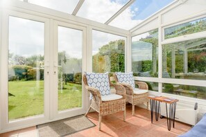 Une véranda avec des fauteuils en osier et une petite table, de grandes portes vitrées et une vue sur un jardin.
