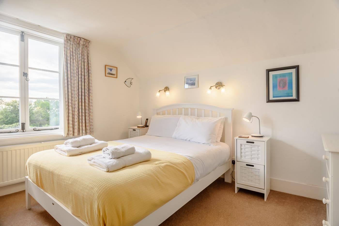 A neatly made bed with white linens and a yellow blanket, a bedside table with a lamp, and a window with curtains.