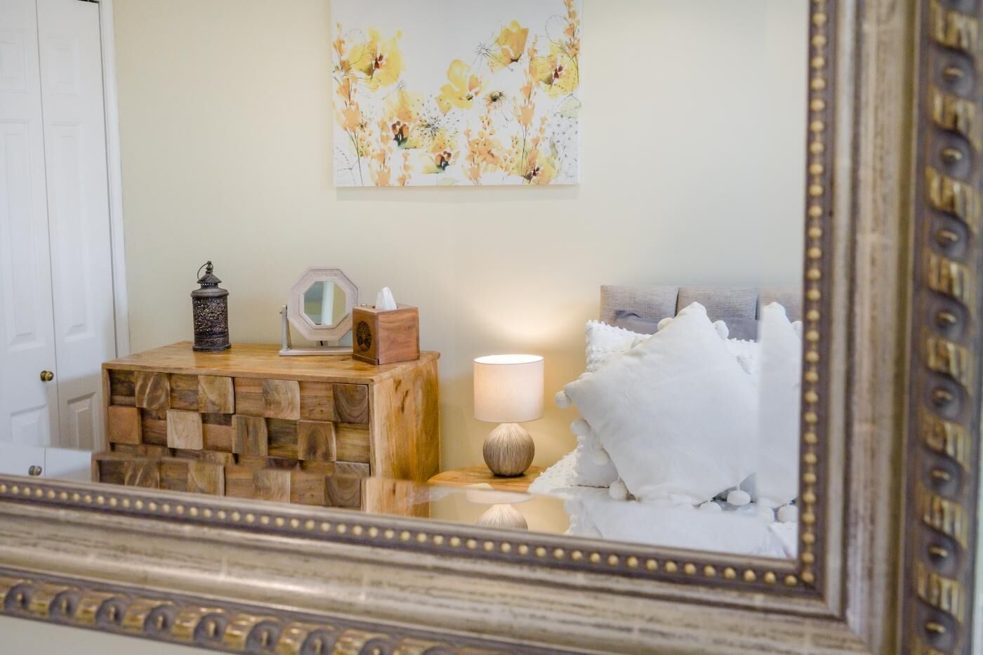 A bedroom with a wooden bedside table, a lamp, a mirror, and a floral artwork on the wall.