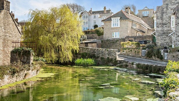 A historic stone building with a small pond, willow trees, and a stone bridge. - 2 Bed in Swanage (oc-wy501) (Swanage)