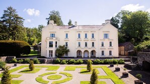 Un grand bâtiment à deux étages, doté d’une façade symétrique, d’une entrée centrale avec un portail en arc de cercle et d’un jardin à la française, caractérisé par des motifs géométriques et des haies taillées.