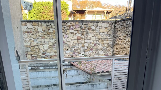 A balcony with a wooden deck, stone wall, and a view of a building with a tiled roof. - Villa Margot (Fumel)