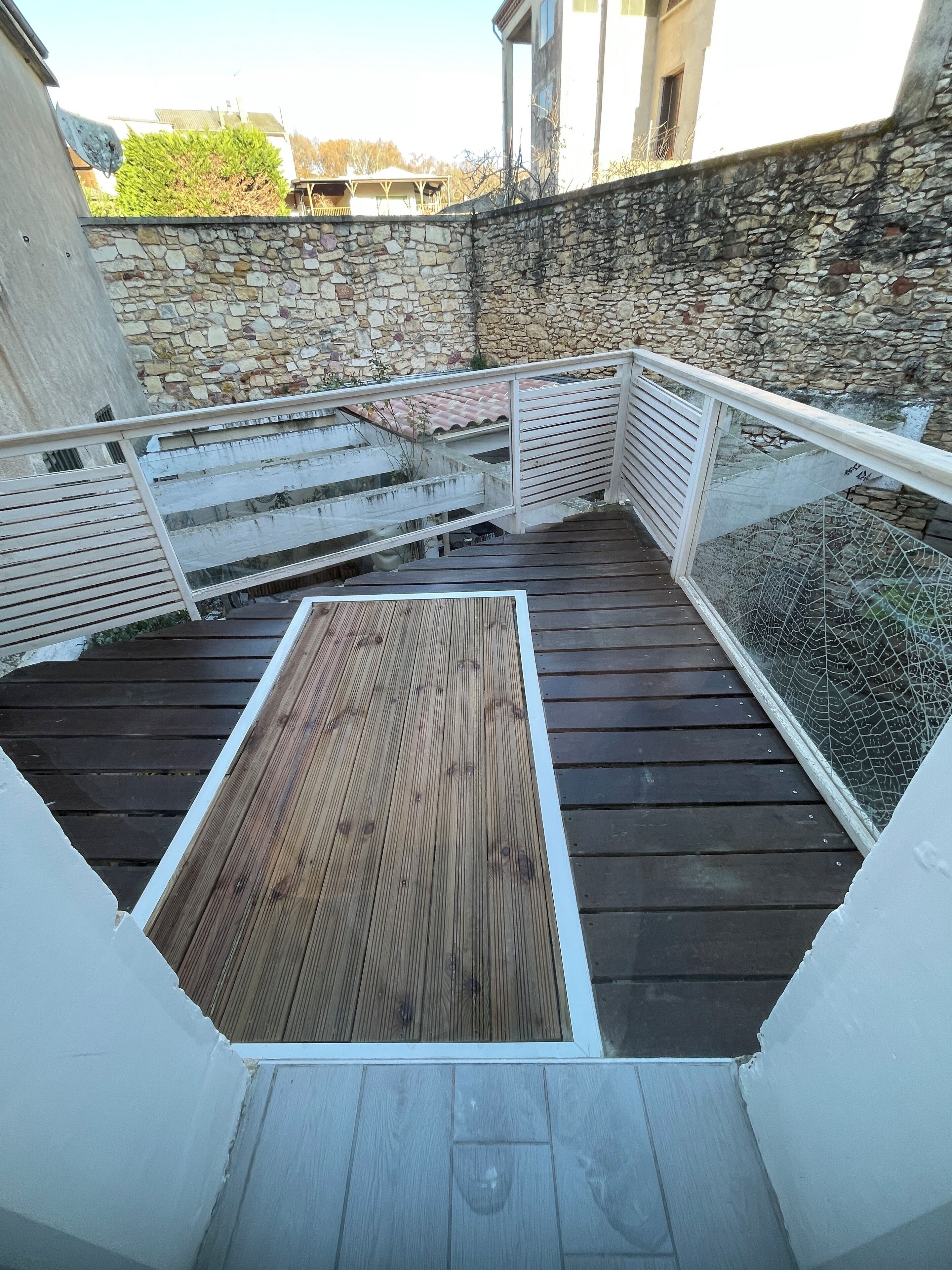 A wooden deck with a white railing, a stone wall, and a view of buildings in the background.