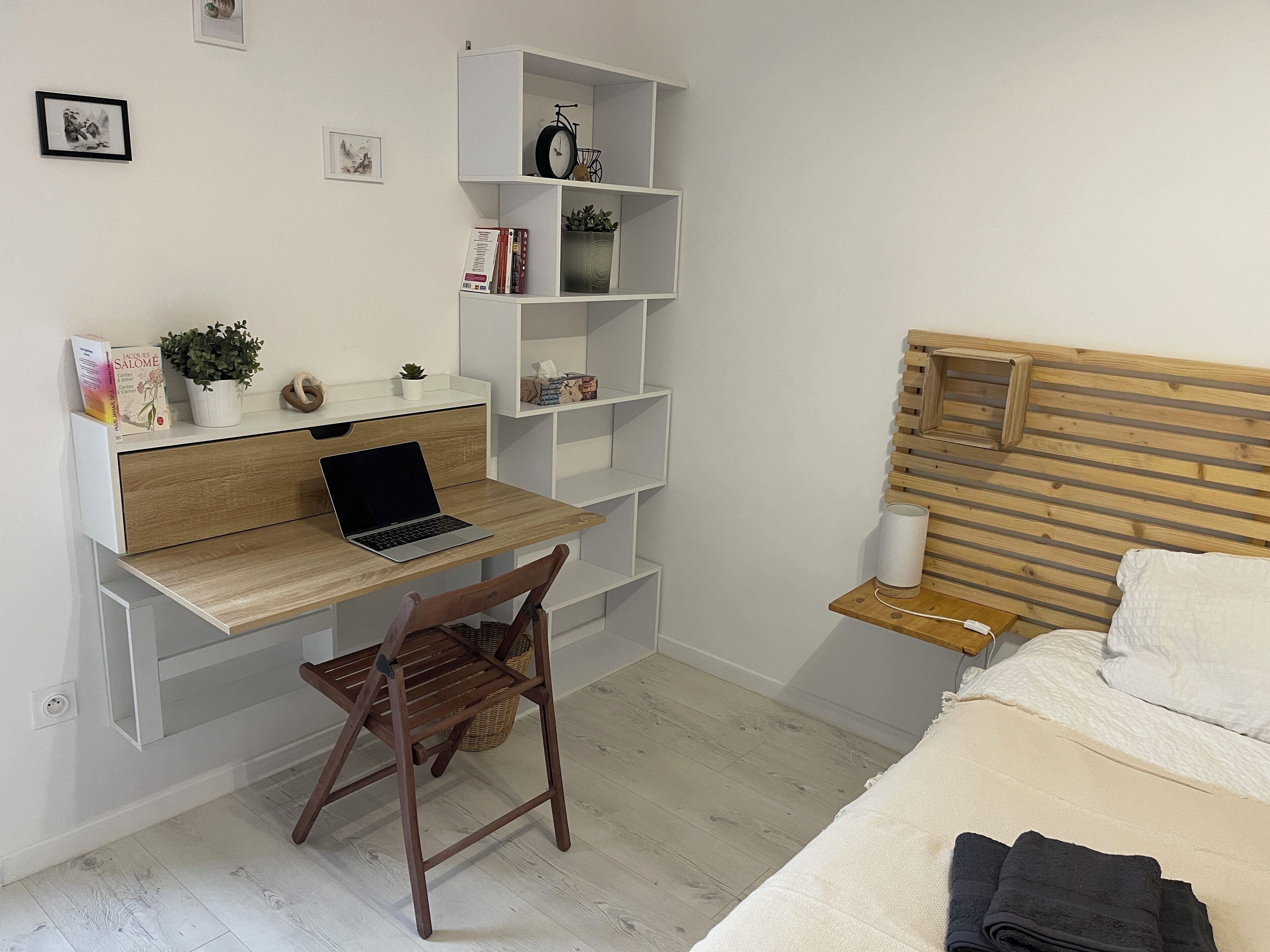 A bedroom with a wooden bed, a desk with a laptop, a chair, a bedside table with a lamp, and shelves with books and a bicycle.