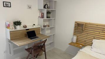 A bedroom with a wooden bed, a desk with a laptop, a chair, a bedside table with a lamp, and shelves with books and a bicycle.