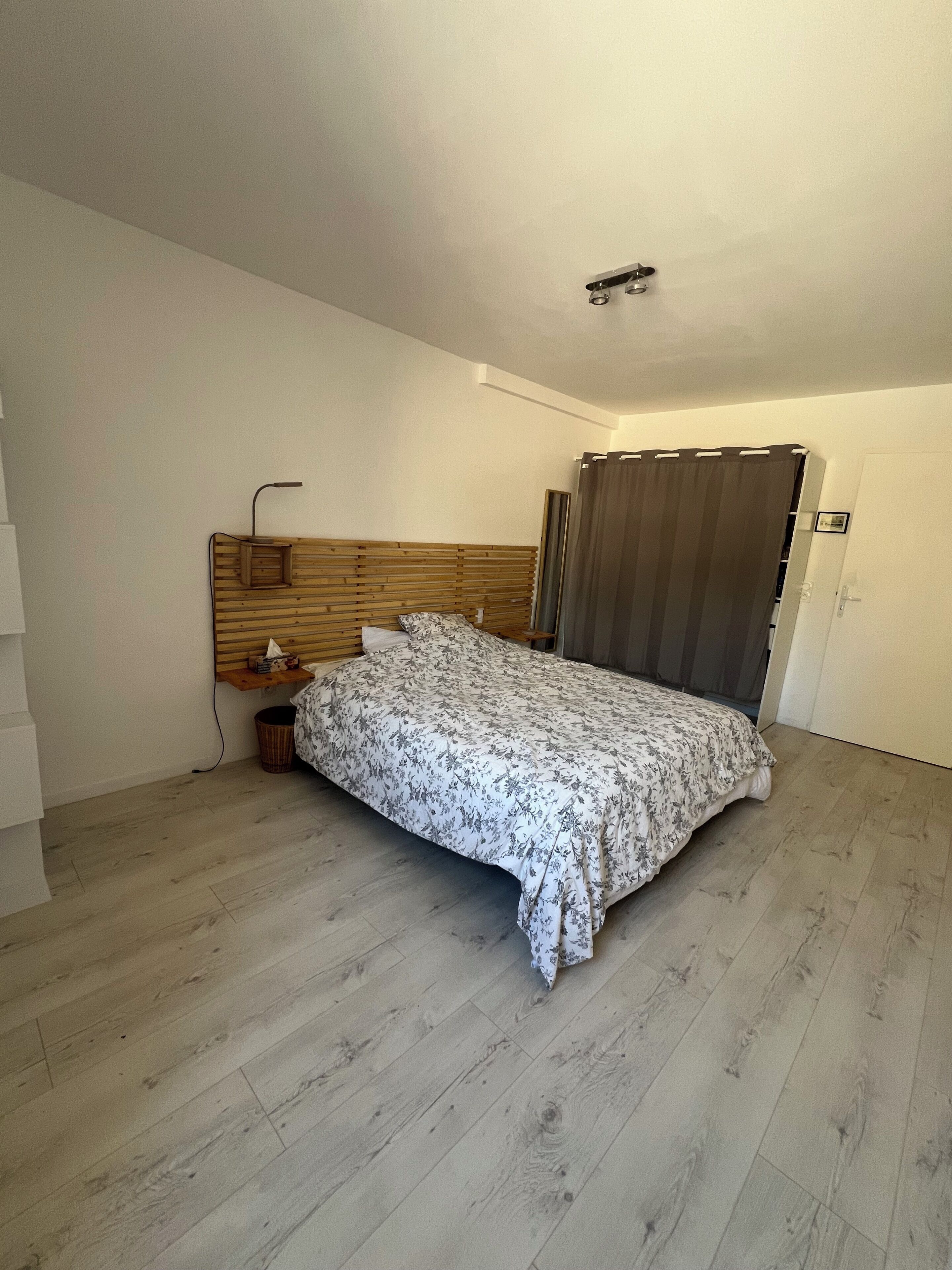 Une chambre avec une tête de lit en bois, un lit recouvert d’une couvre-lit à motifs floraux, une table de chevet avec une lampe et un rideau à la fenêtre.