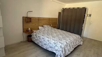 Une chambre avec une tête de lit en bois, un lit recouvert d’une couvre-lit à motifs floraux, une table de chevet avec une lampe et un rideau à la fenêtre.
