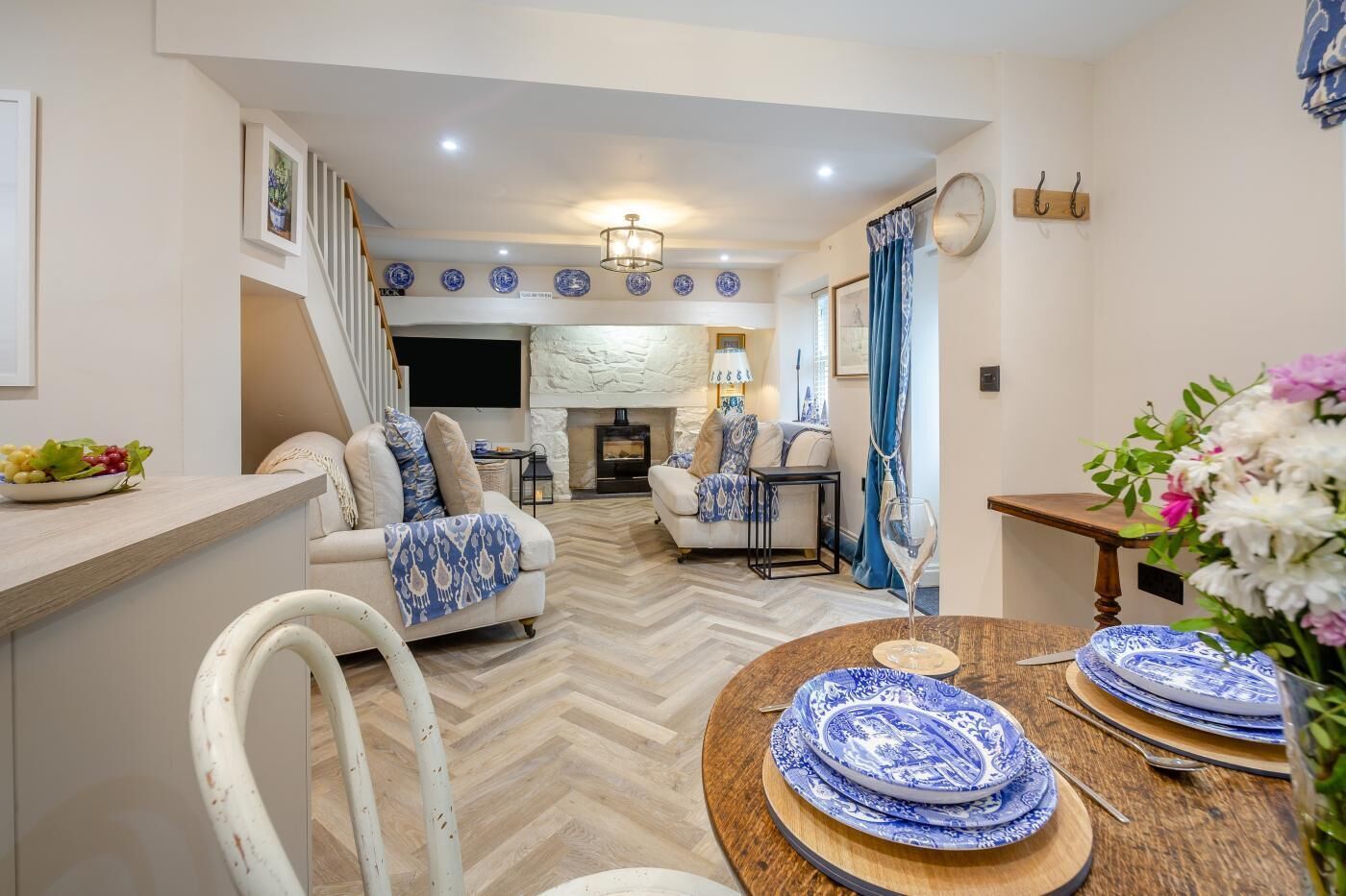 Un salon moderne avec une cheminée, un coin repas avec de l’argenterie bleue et blanche à motifs, et un parquet en bois disposé en chevrons.