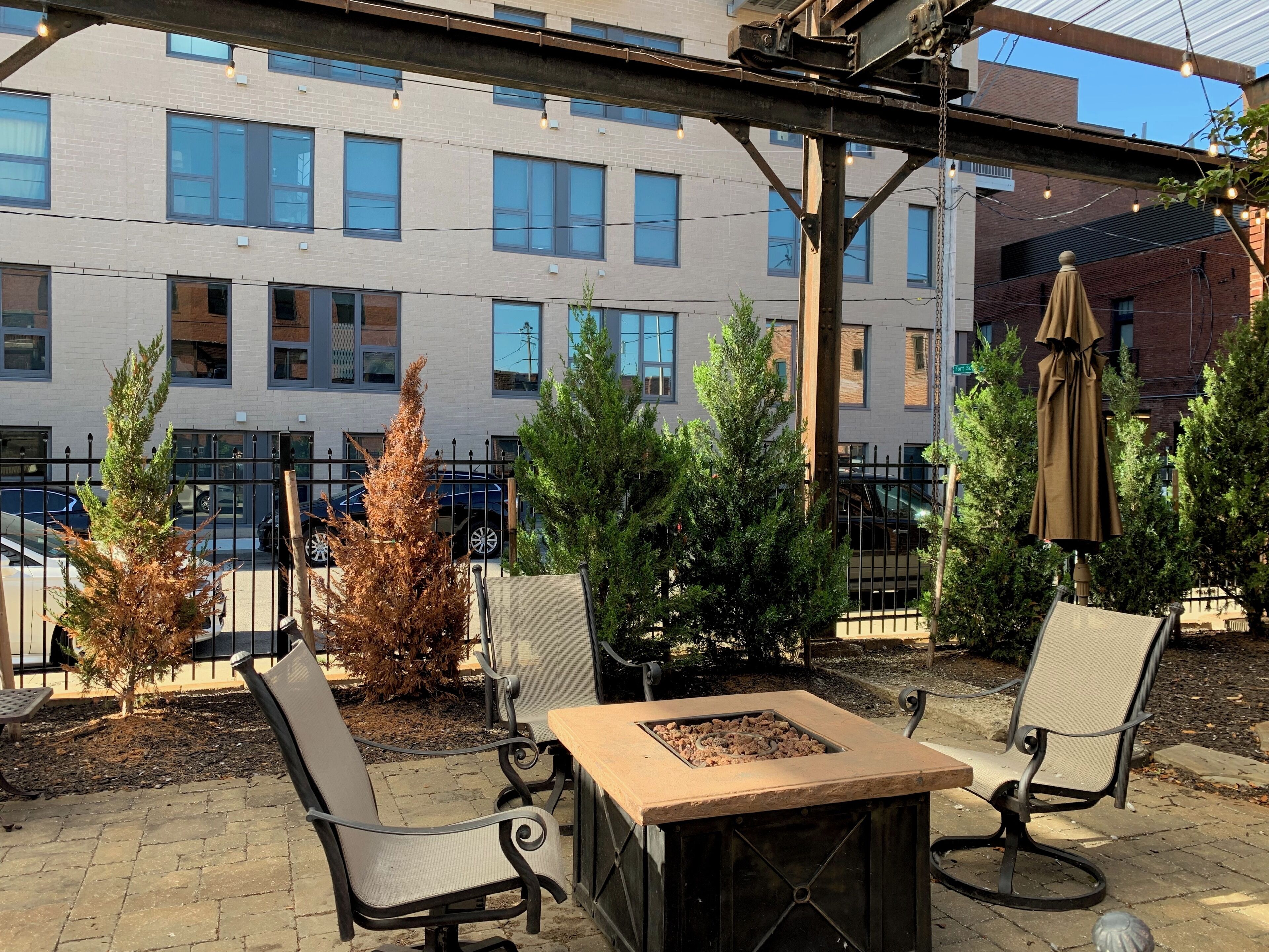 An outdoor patio with seating and a fire pit, surrounded by trees and a building in the background.