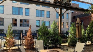 Patio esterno con posti a sedere e un braciere, immerso tra alberi e con un edificio sullo sfondo.