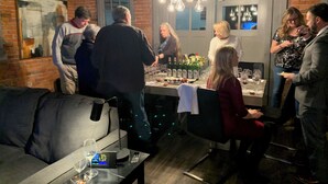 Un gruppo di persone in una stanza con pareti in mattoni, lampade a sospensione, un tavolo con bicchieri e bottiglie di vino e un divano con un telecomando sopra.
