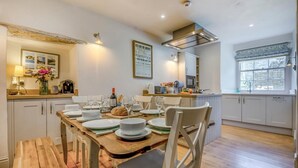 A dining area with a wooden table set for four, white chairs, a kitchen with white cabinets, and a window with blinds. - 3 Bed in Swanage (oc-wy557) (Swanage)