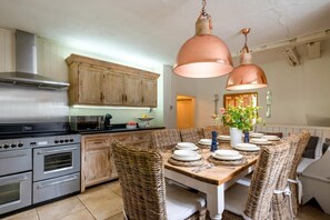 A kitchen with a dining table set for four, surrounded by wicker chairs, a stainless steel stove, and copper pendant lights. - 5 Bed in Halesworth (oc-cfl) (Halesworth)