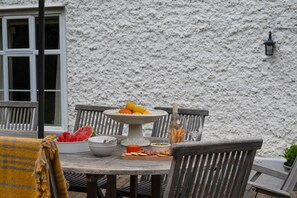 A round wooden table with a white plate of fruit, a bowl of watermelon, and a bottle of wine, set against a textured white wall with a window and a lamp. - 5 Bed in Halesworth (oc-cfl) (Halesworth)