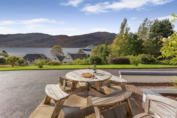 A wooden outdoor dining area with a table and benches, overlooking a lake and houses in the background. - 3 Bed in Ullapool (oc-s33063) (Ullapool)