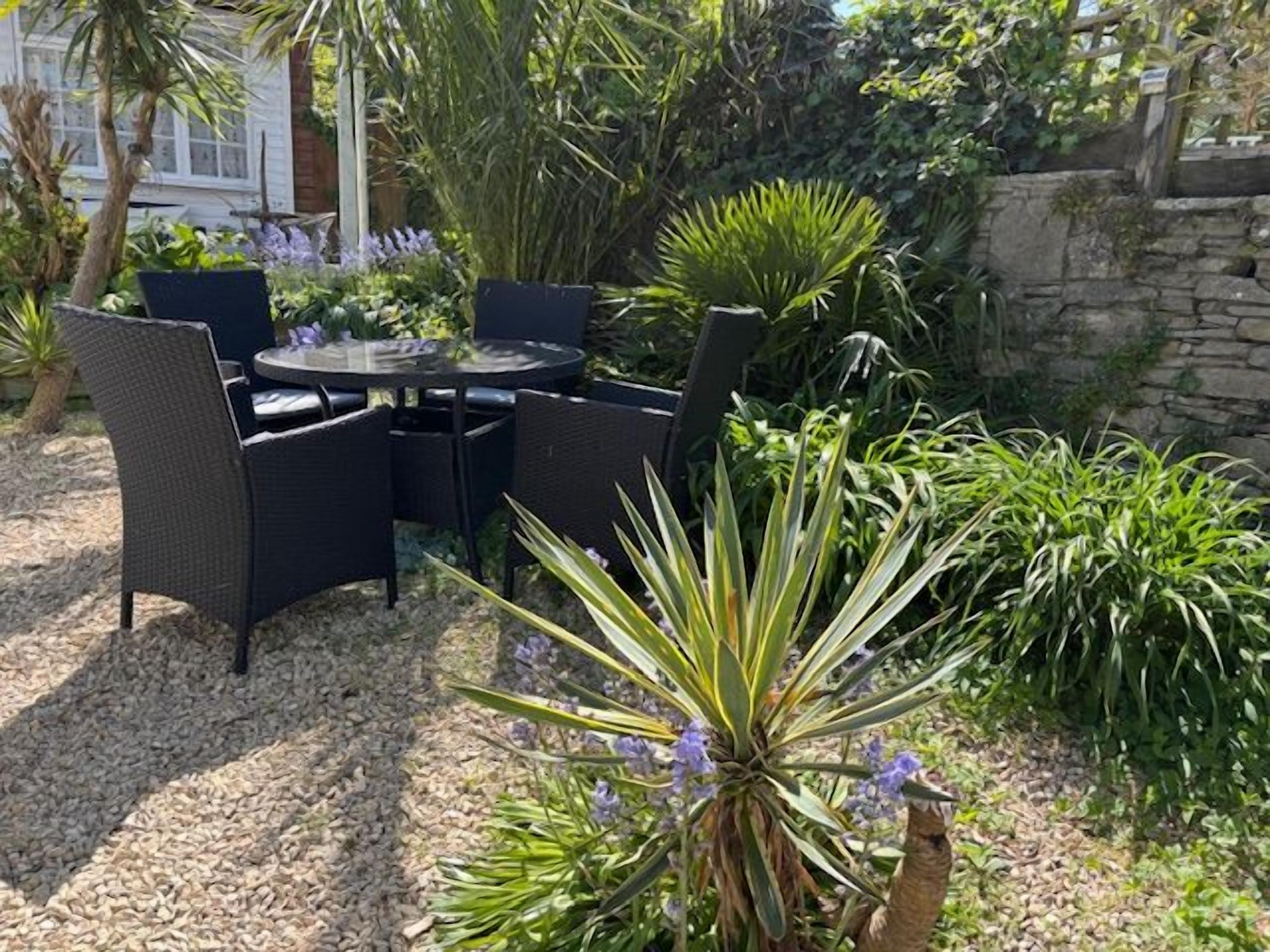 Un patio exterior con una mesa redonda y sillas, rodeado de plantas y un muro de piedra.