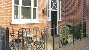 A brick building with a white-framed window, a door with a decorative glass panel, and a small outdoor seating area with metal chairs and a table. - 2 Bed in Southwold (oc-sl135) (Southwold)