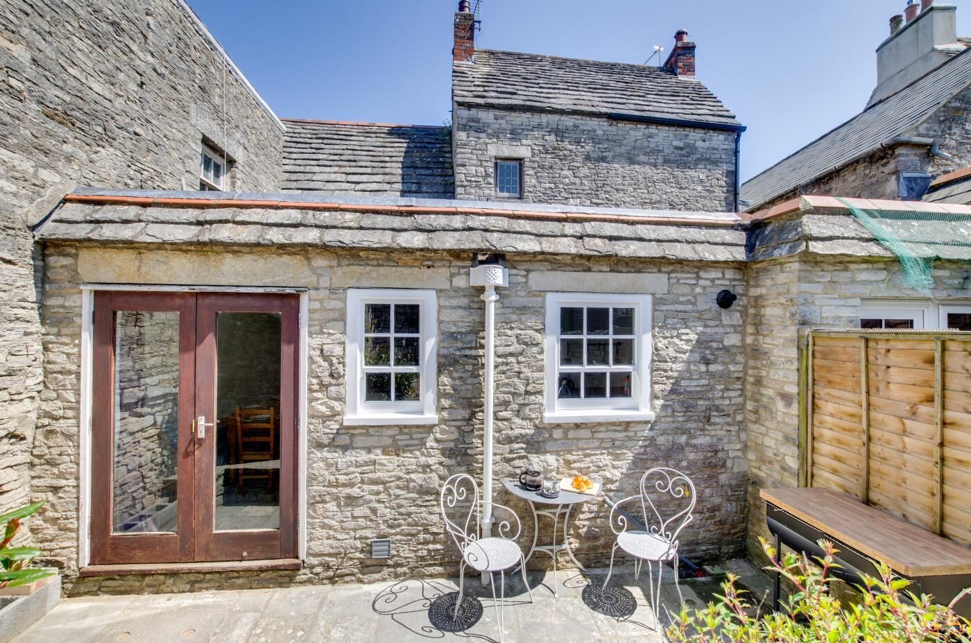 A stone cottage with a wooden door, two windows, and a small patio with metal chairs and a table.