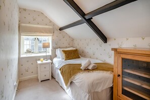 Ein Schlafzimmer mit schrägen Wänden, einem Fenster mit Aussicht, einem Nachttisch, einem Bett mit Kopfteil und einem Holzkasten.