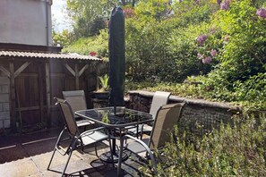 Une terrasse extérieure avec une table et des chaises en verre, entourée d’une végétation luxuriante et d’un mur de pierre.