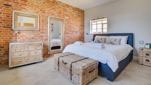 Un dormitorio con pared de ladrillo, una cama con ropa de cama blanca, un aparador, un espejo y una ventana.