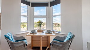 Een zithoek met een tafel voor twee personen, twee rieten stoelen, uitzicht op een stad door grote ramen, en een kleine vaas met bloemen op de tafel.