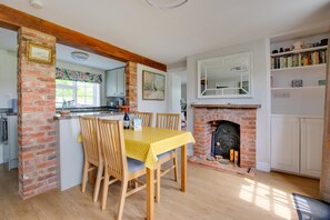 A dining area with a brick fireplace, wooden chairs, a yellow tablecloth, and a kitchen in the background. - 2 Bed in Saxmundham (oc-rvr) (Snape)