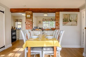Un coin repas avec une nappe jaune, des chaises blanches, un mur en briques et une cuisine en arrière-plan.