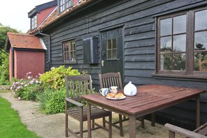 Une table en bois avec une chaise, une assiette contenant des croissants, une théière et une tasse, le tout placé à l’extérieur d’une maison avec une porte rouge et une fenêtre.