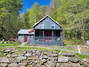 A two-story house with a porch, a red door, and a stone wall in front.