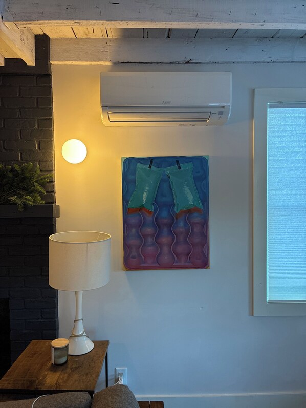 A room with a brick wall, a white air conditioning unit, a round wall light, a table with a lamp and a candle, and a colorful artwork.