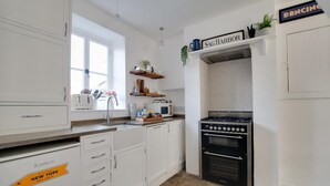 Cocina compacta con muebles blancos, estufa a gas, microondas y pileta. Hay objetos decorativos en los estantes y una ventana que proporciona luz natural.