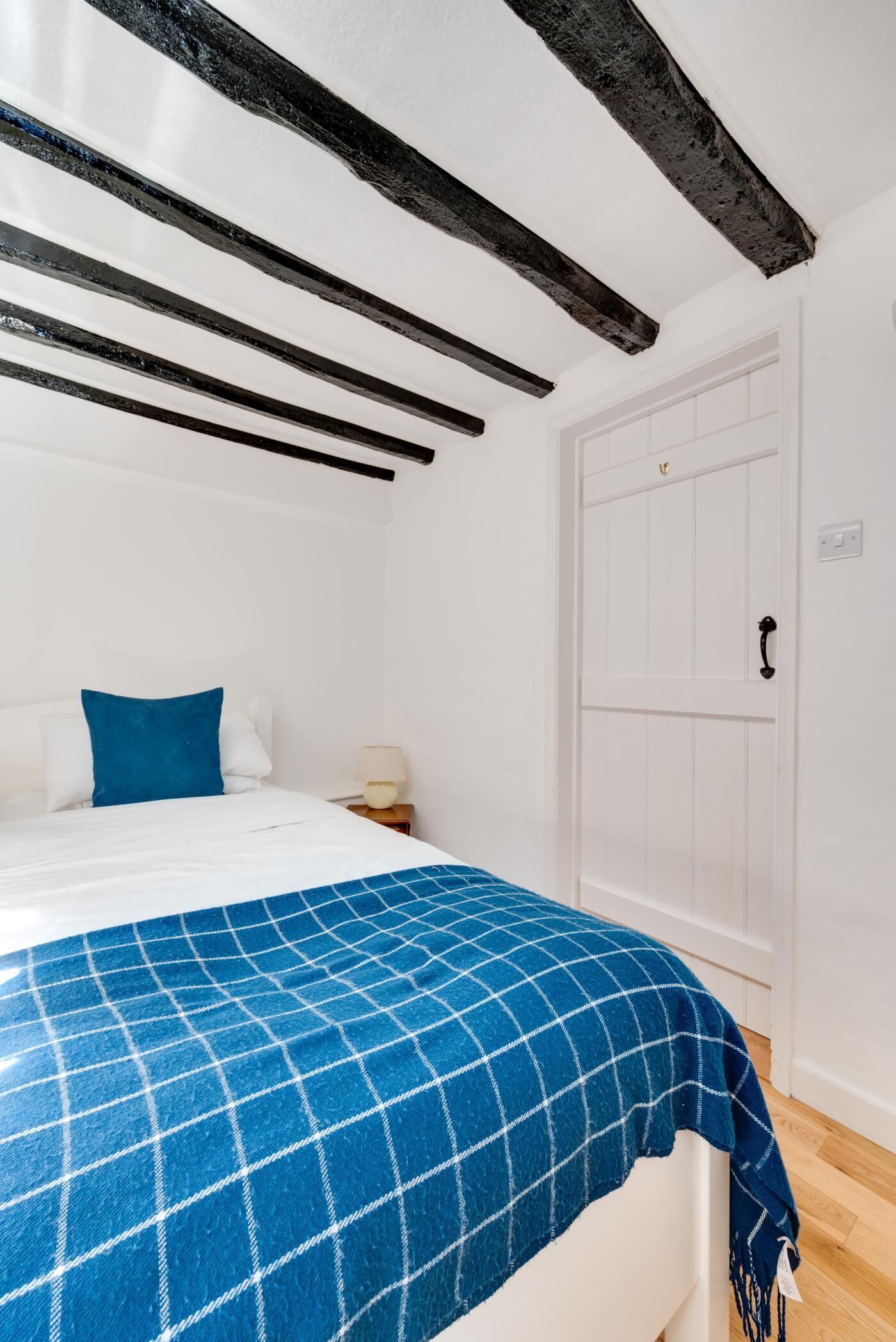 A bedroom with a bed, a blue checkered blanket, a white door, and exposed wooden beams on the ceiling.