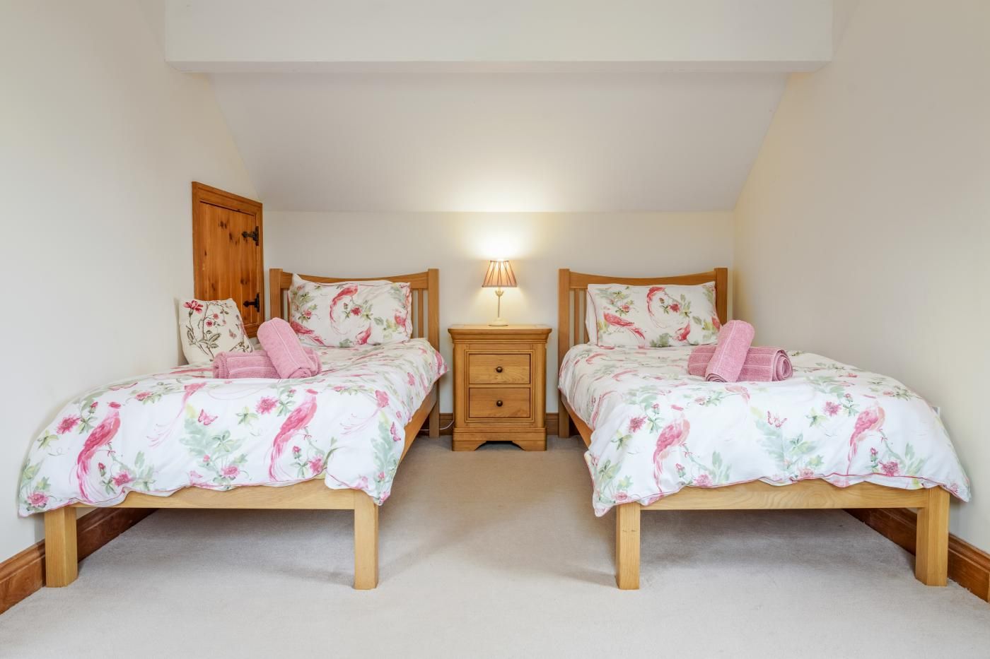 A bedroom with two wooden beds, each with floral bedding and pink towels, a bedside table with a lamp, and a wooden wardrobe.