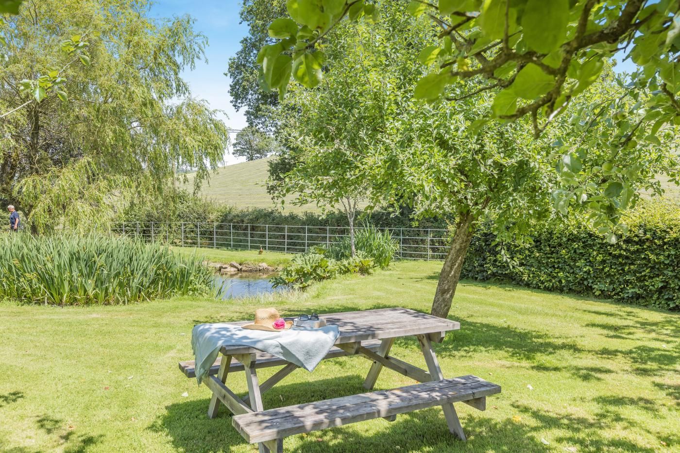 Ein Picknicktisch mit Tischdecke und Hut, umgeben von üppigem Grün und einem kleinen Teich.